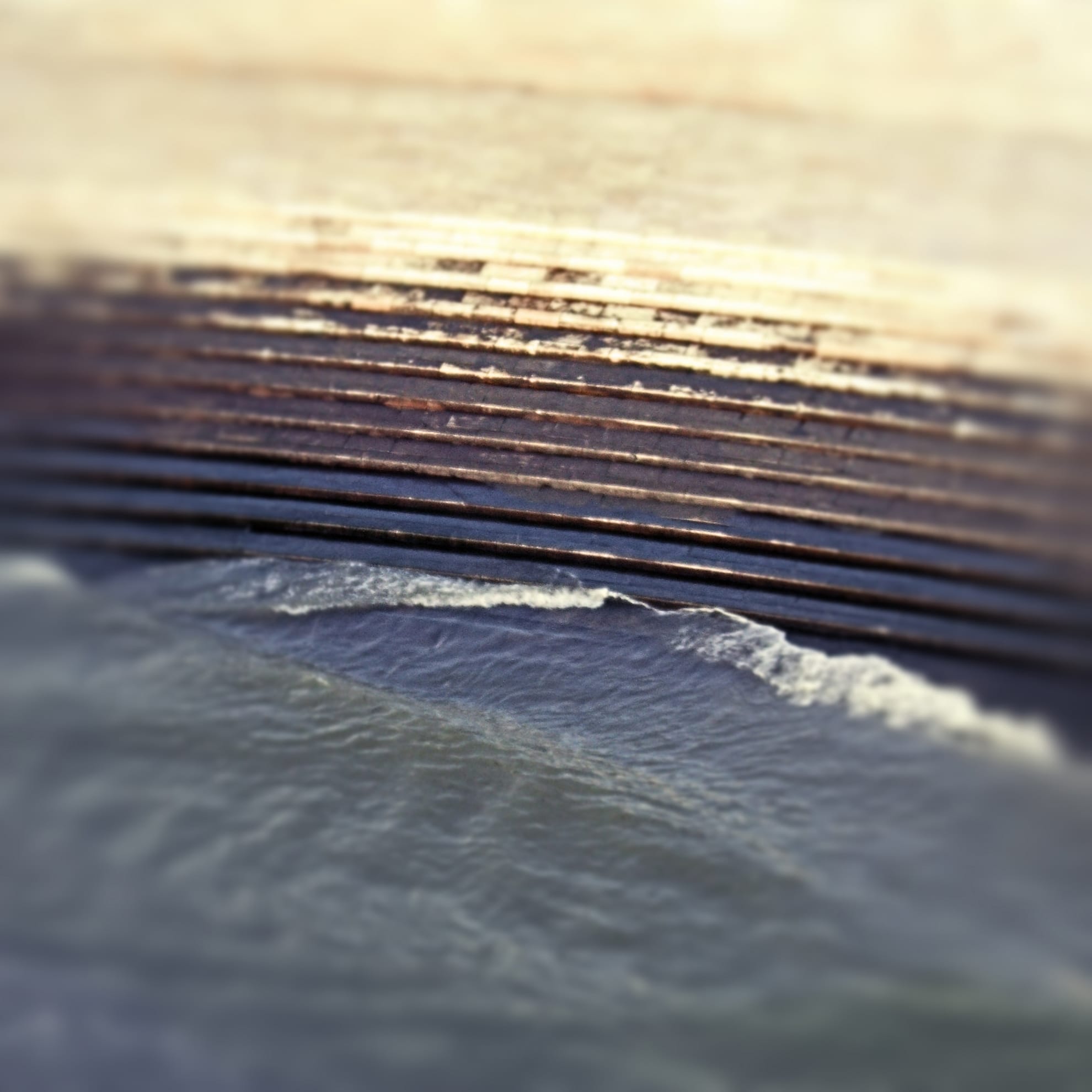 A bird's-eye view shows ocean waves gently crashing against a multi-layered, stone seawall with a blurred effect creating an ethereal, serene ambiance. The layers of the seawall have varying shades of beige, resembling the ancient majesty of Belem Tower, while the water has a soft gradient mix of blue and grey tones.