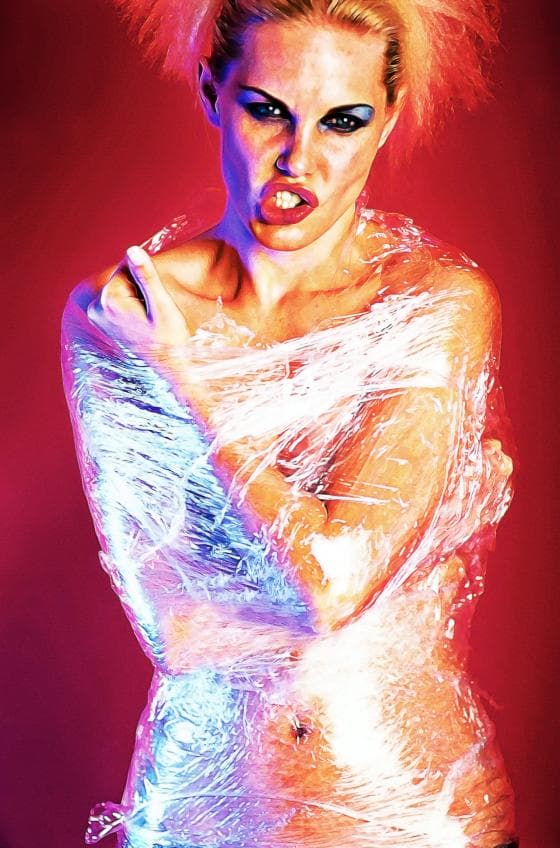 A person with vivid makeup and a serious expression is wrapped in plastic film, covering the body from shoulders to hips. Born Into Wrap, the background is a vibrant red, contrasting with the soft lighting on the subject. The hair is styled with voluminous, fluffy, pink accents.