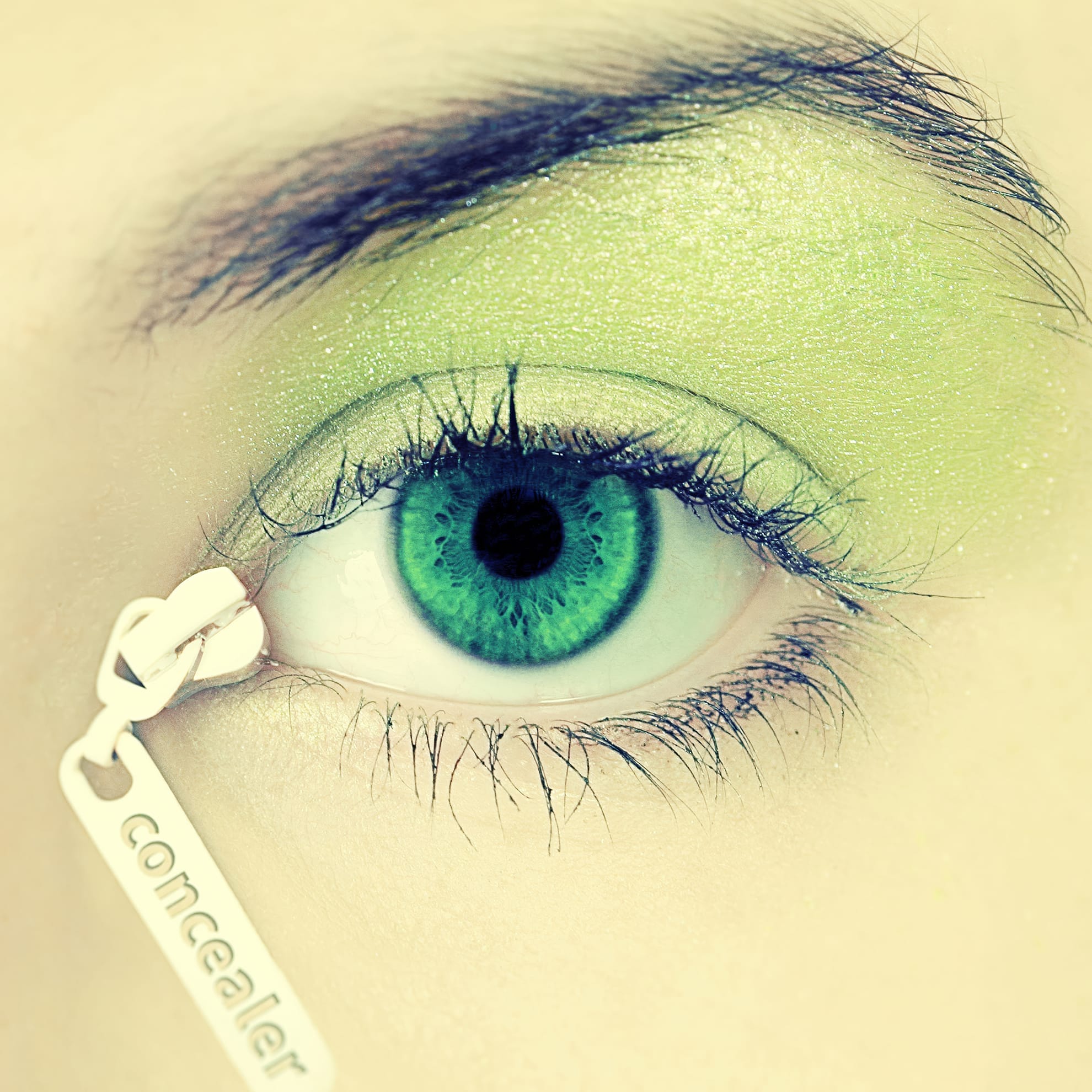 A close-up of an eye with a green iris and green eyeshadow. The defined lashes accentuate the beauty, while a white tag with the word "concealer" written on it hangs from the lower eyelid, creating a surreal and artistic effect. The overall tone of the image is cool, blending green and white hues.
