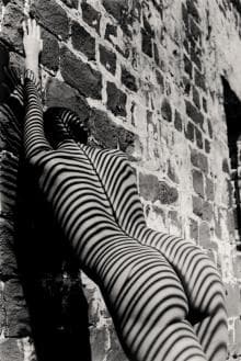 A person stands against a textured brick wall, with their back to the camera. They are covered in striped shadows projecting across their figure, creating a pattern of dark and light lines over the entire body. The composition includes strong contrasts and artistic lighting.