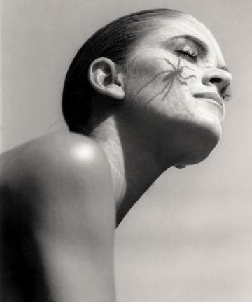 A grayscale photo shows a person with closed eyes, head tilted back, and a small spider on their cheek. The lighting casts shadows, highlighting smooth skin and long eyelashes. The background is a clear sky, giving a serene and contemplative mood.