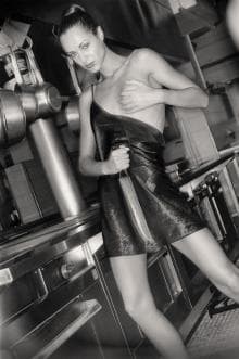 A black-and-white image of a person standing in a kitchen, holding a large knife. They wear a short, sleeveless leather outfit and pose with one hand on their chest. The background features metallic kitchen appliances and fixtures, enhancing the industrial feel.