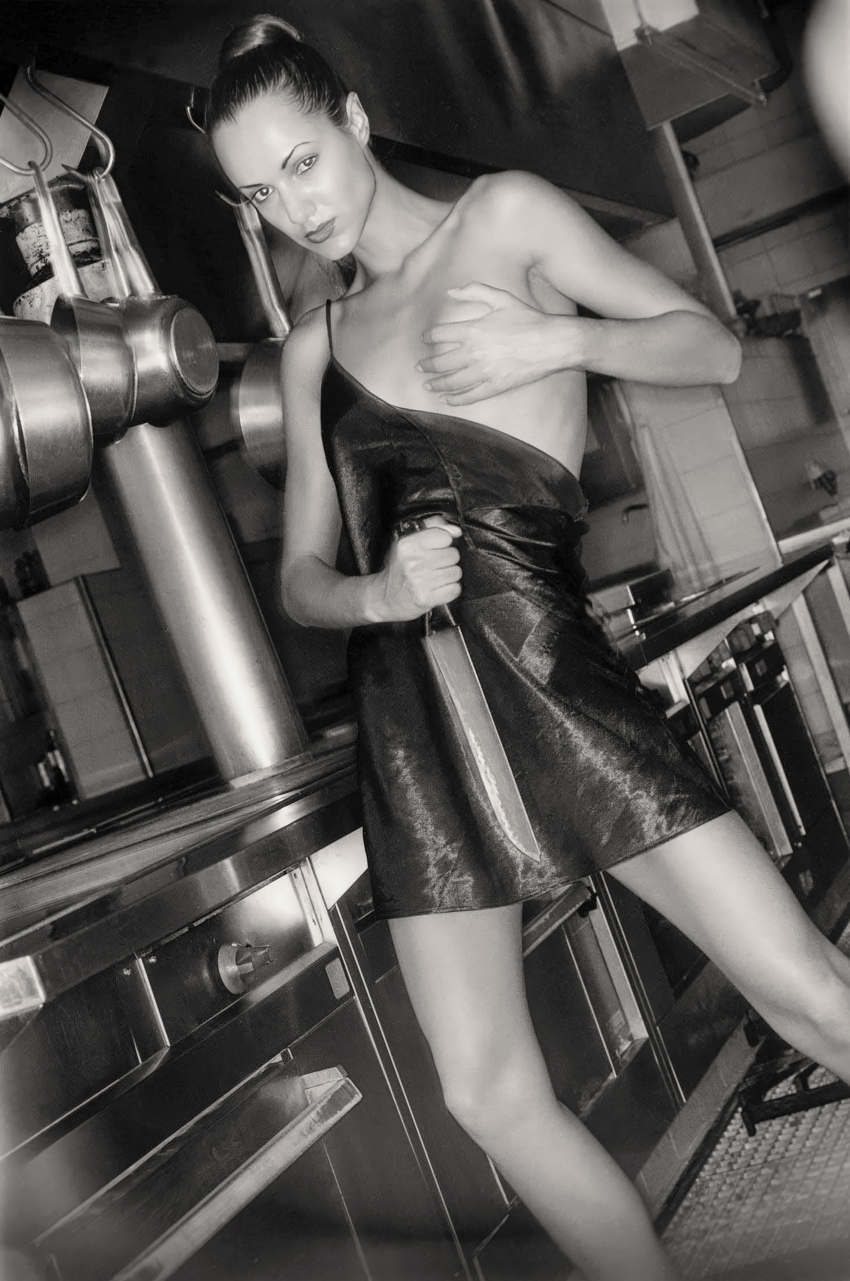 A black-and-white image of a person standing in a kitchen, holding a large knife. They wear a short, sleeveless leather outfit and pose with one hand on their chest. The background features metallic kitchen appliances and fixtures, enhancing the industrial feel.