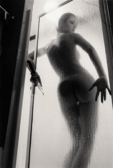 Silhouette of a person behind a frosted glass shower door, creating a blurred effect. The person has one hand on the door handle and the other against the glass, with light casting shadows. The scene suggests privacy and mystery.