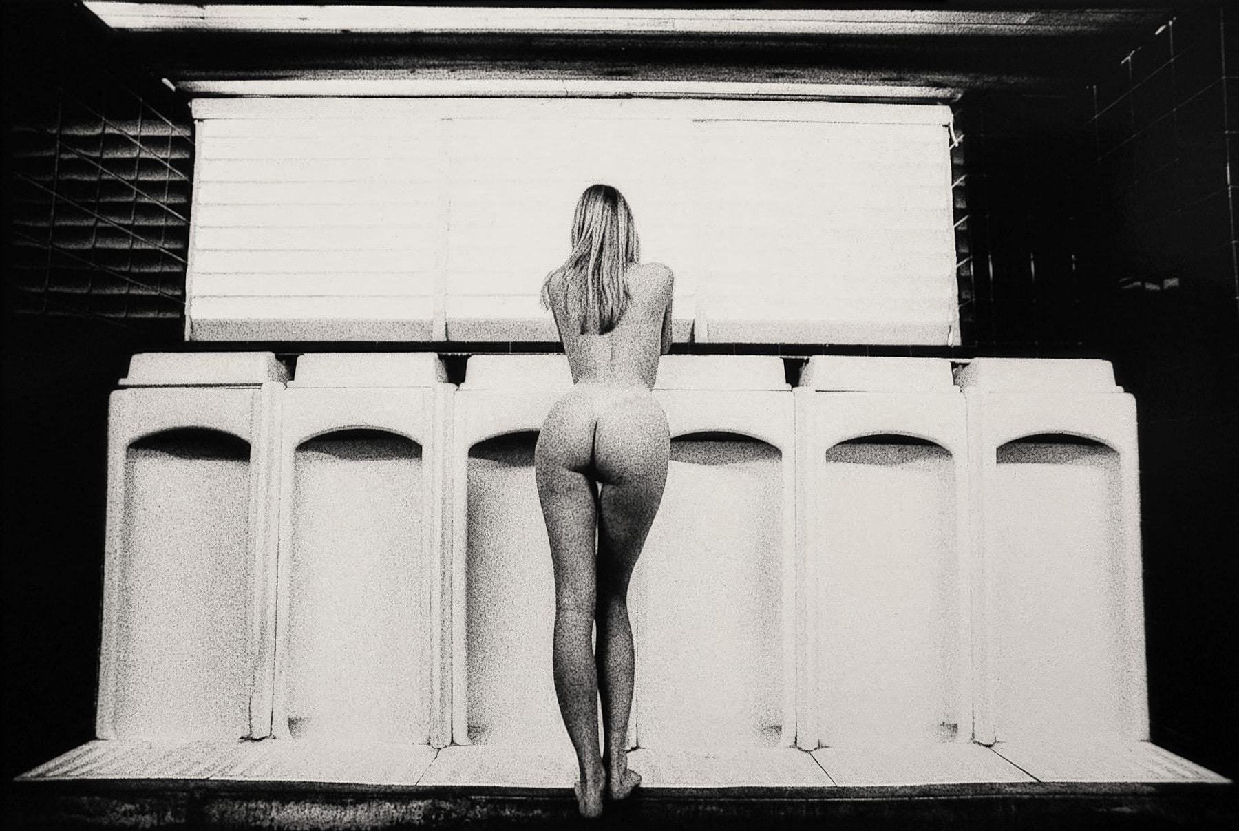 A black-and-white photo shows a nude person with long hair, standing at a row of urinals, facing away from the camera. The setting has a tiled wall and a large window above the urinals. The lighting and composition create a stark, artistic contrast.