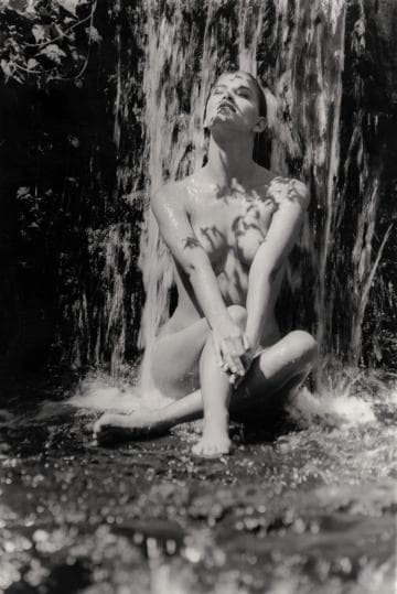 A nude woman sits cross-legged under a waterfall, gazing upwards with her arms crossed over her knees. Sunlight casts leaf shadows on her skin and the flowing water. The scene conveys a sense of tranquility and nature's embrace.