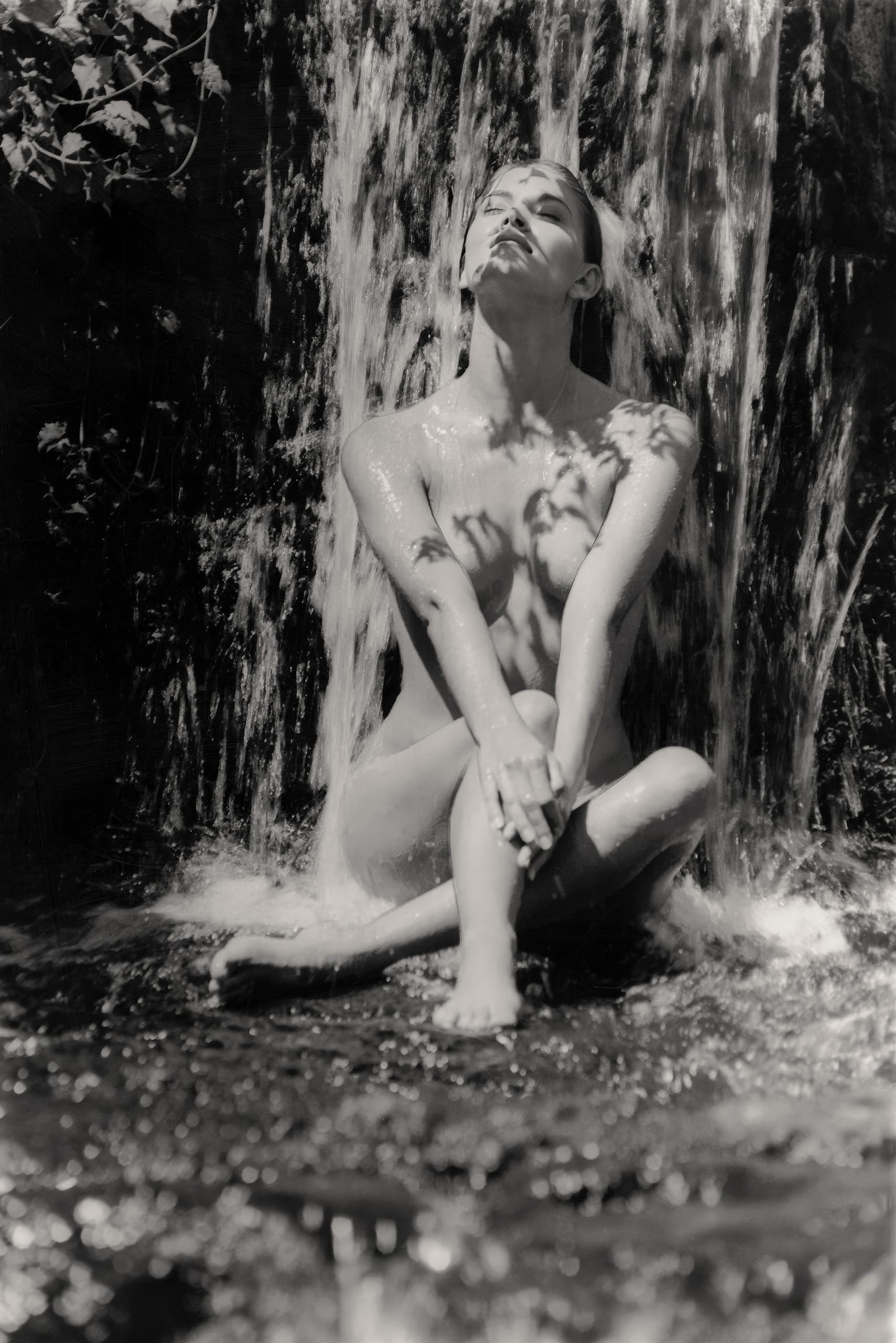A nude woman sits cross-legged under a waterfall, gazing upwards with her arms crossed over her knees. Sunlight casts leaf shadows on her skin and the flowing water. The scene conveys a sense of tranquility and nature's embrace.