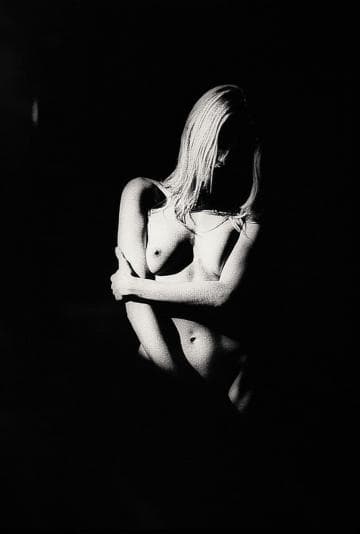 A black and white photograph of a nude person with long hair. They are sitting with their arms crossed over their knees, partially obscured. The image is dramatically lit, casting deep shadows and highlighting the contours of their body, creating a moody, artistic effect.