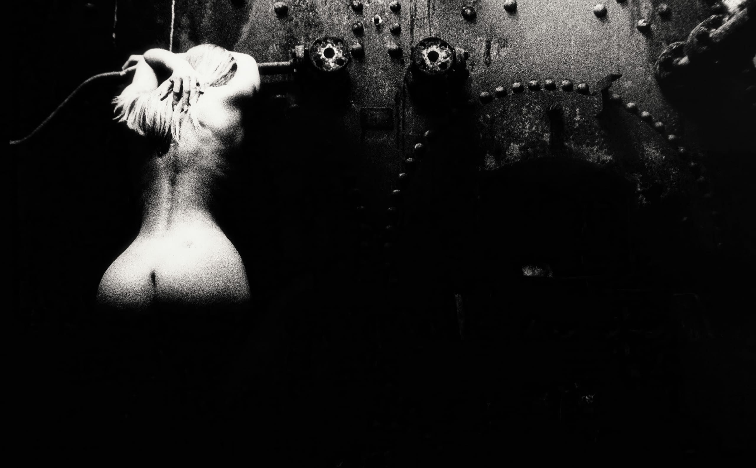 A black and white image shows a naked person with long hair facing a dark, industrial wall. They have their arms raised, leaning against the wall. The scene is dimly lit, emphasizing shadows and creating a dramatic, moody atmosphere. Industrial elements like bolts and gears are visible.
