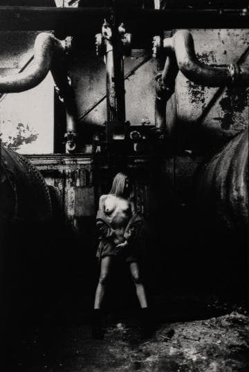 A black-and-white photograph shows a person standing in an industrial setting. They are in front of large pipes and machinery, partially unclothed, with lighting casting deep shadows. The area looks worn and gritty, contributing to a dramatic and edgy atmosphere.