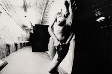 Black and white image of a nude person standing in a narrow, dimly lit hallway with tiled walls and a wooden ceiling. The person, partially obscured by shadows, holds a gun by their hip. The perspective is tilted, adding a dramatic and intense atmosphere to the scene.