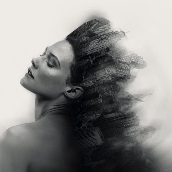 A black and white surrealistic image shows a woman's serene face and bare shoulders blending seamlessly into a skyline of tall buildings. The cityscape appears to emerge like a crown from her head, creating a fusion of human and urban elements, evoking a dreamlike, introspective atmosphere.