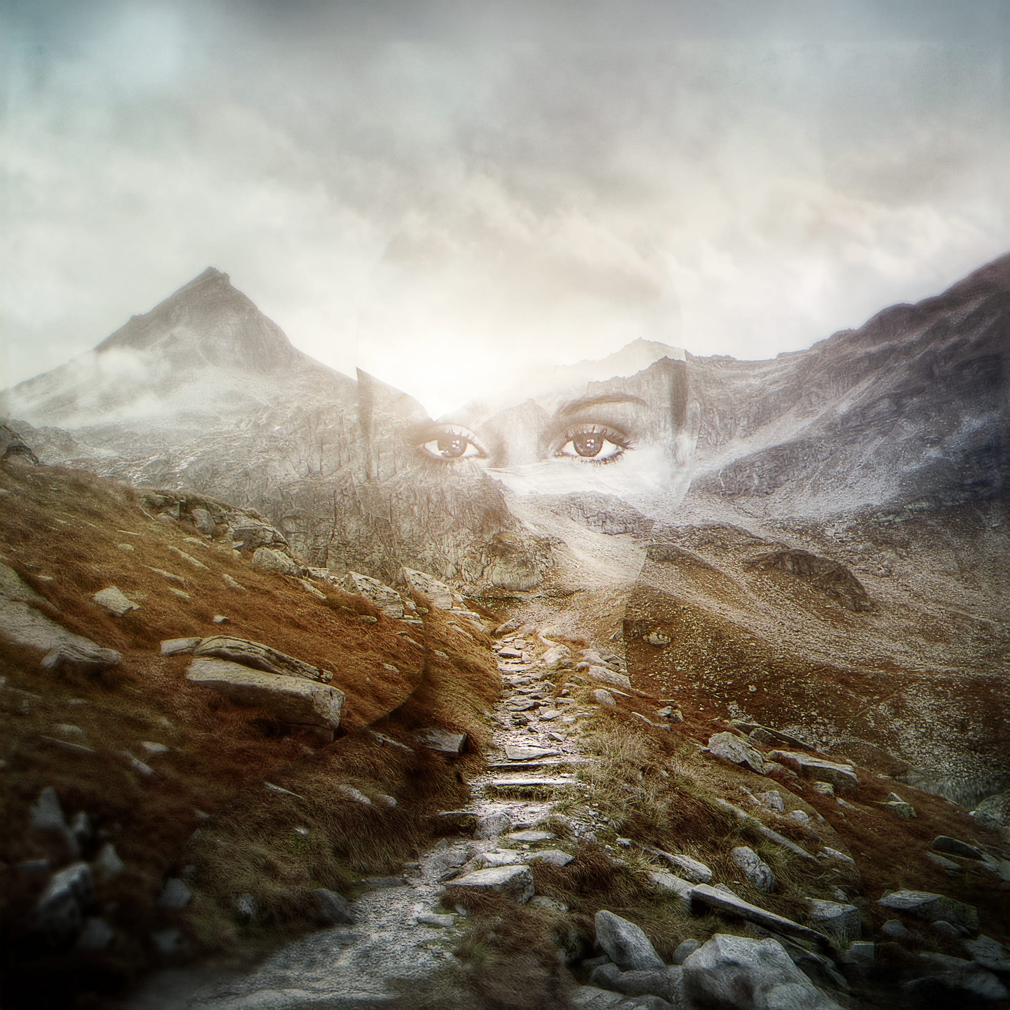 A surreal landscape features a rocky mountain path beneath a cloudy sky. Overlaid on the scene is a translucent image of a person's eyes, blending seamlessly with the terrain, creating a mystical and ethereal atmosphere. The overall tone is moody and contemplative.
