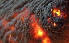 Close-up of glowing embers nestled within the textured ridges of a wood surface, creating a vivid, fiery effect. The embers radiate orange and yellow light, contrasting against the darker, charred wood, evoking a sense of warmth and intensity.