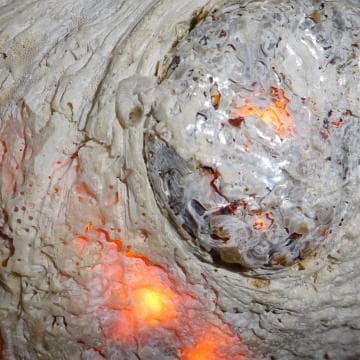 Close-up of a textured surface resembling coral or rock, with intricate patterns and crevices. Bright, warm orange lights glow from within, casting a fiery reflection. The overall appearance suggests a blend of natural and abstract elements, merging the organic with the luminous.