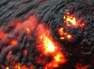 Close-up of glowing embers in wood, showing orange and red hues beneath the charred, ashy surface. The fiery textures create a swirling pattern, with bright spots of light peeking through darker areas, evoking a sense of intense heat and dynamic movement.