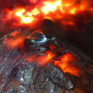Close-up of molten lava, showing a glowing orange and red surface with fiery reflections. The texture appears fluid and dynamic, with dark, metallic-looking areas cooling over the lava flow. Bright, intense colors create a dramatic and fiery scene, suggesting a volcanic environment.