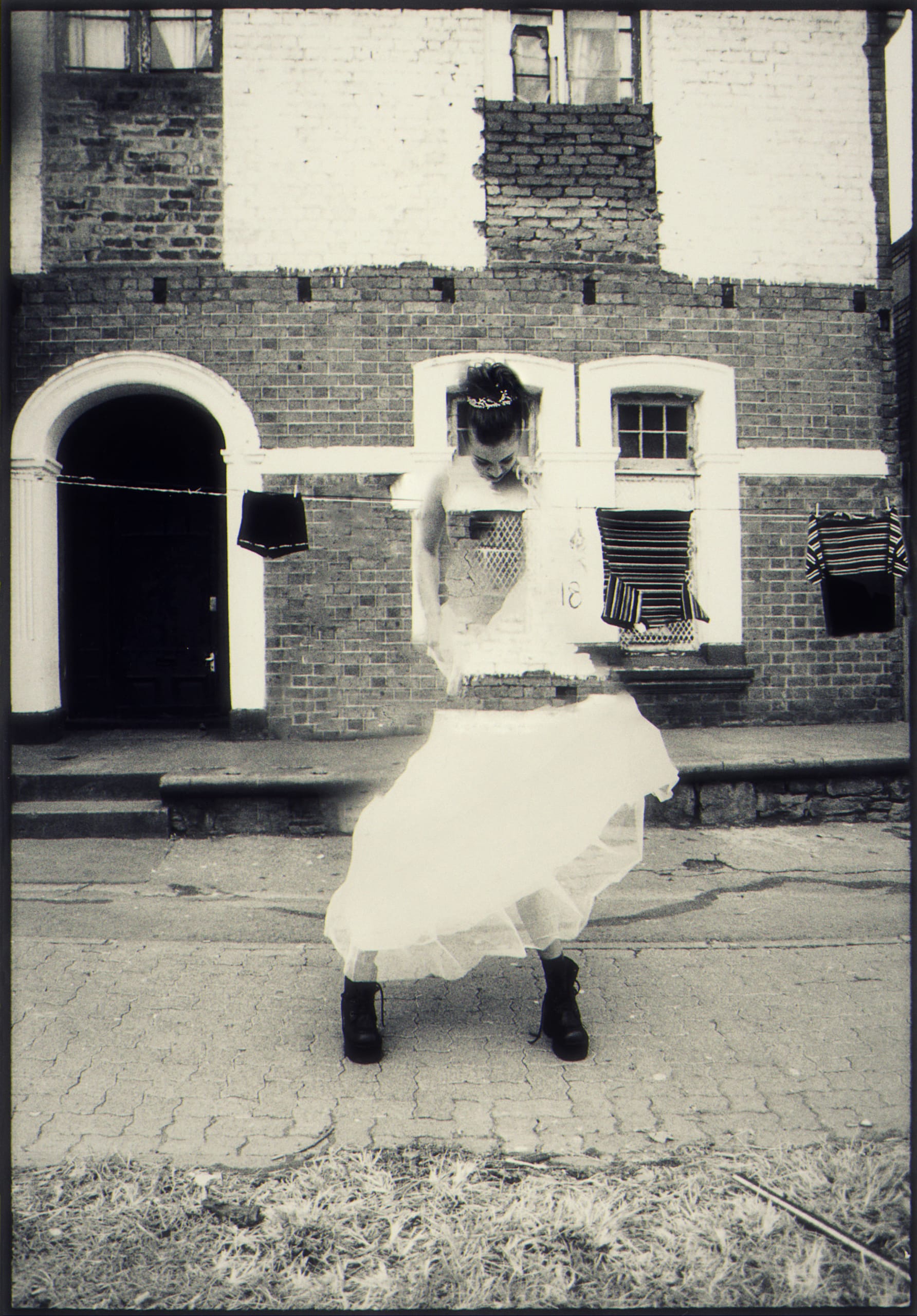 Black and white photo of a person in a white dress and boots, standing in front of a brick building with arched doorways and windows. They hold the hem of their skirt. Clothes, including shirts, hang on a line nearby. The ground is cracked, and grass grows in the foreground.