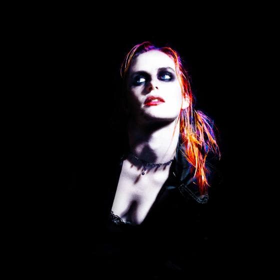A woman with vibrant red hair is illuminated against a dark background. She’s wearing dark eye makeup and red lipstick, and her expression is intense. Her outfit is black, and a necklace is visible around her neck. The lighting creates dramatic shadows on her face.