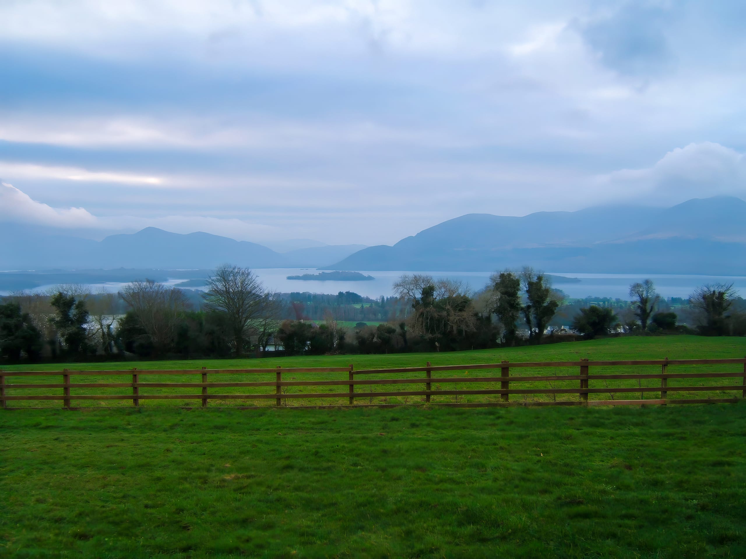A serene landscape features a wooden fence in the foreground, with a lush green field beyond it. Trees line the middle ground, and a tranquil lake with shimmering water and small islands is set against a backdrop of misty mountains under a cloudy sky. The overall atmosphere is peaceful and picturesque, showcasing the beauty of nature.