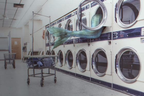 In a bustling laundromat scene with rows of white dryers and washing machines, a mermaid tail humorously extends out of one dryer. In the foreground, an empty laundry cart with a few pieces of clothing inside sits on the floor, adding to the whimsical twist on everyday laundry services.