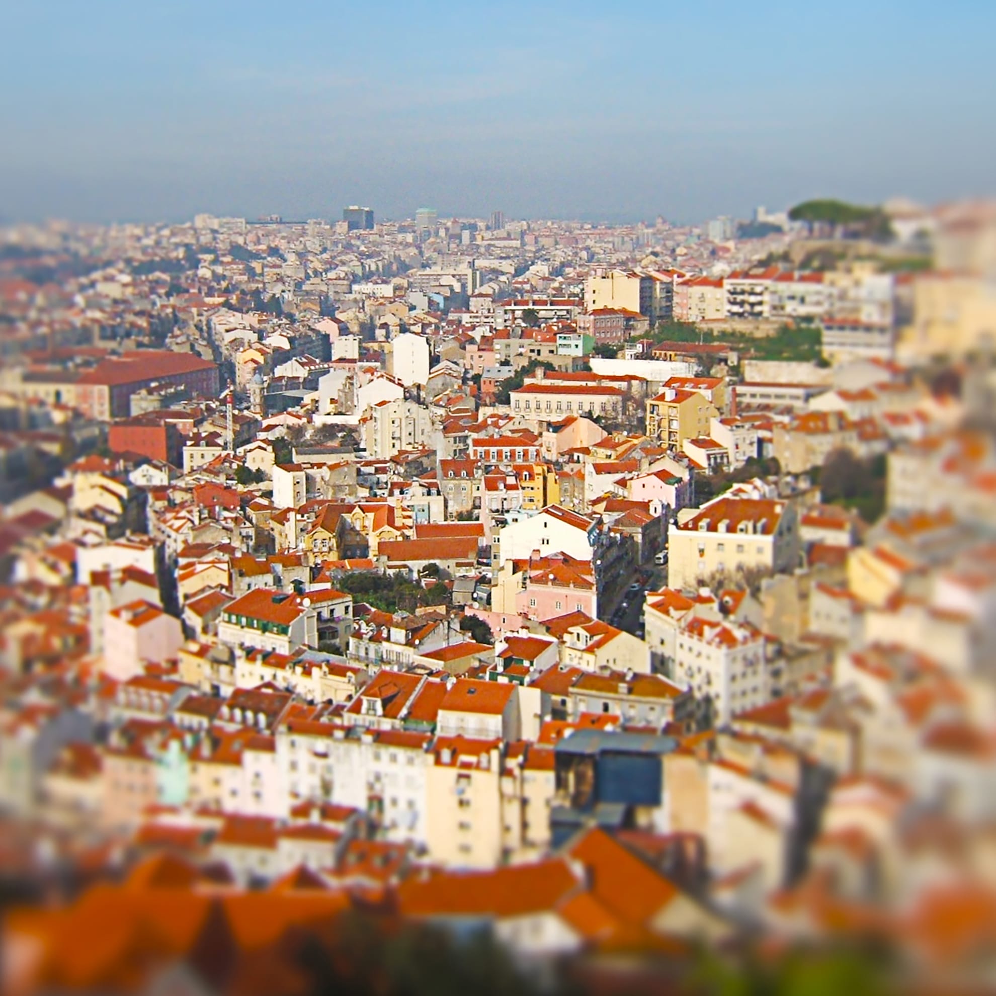 A panoramic view of Lisbon's cityscape featuring densely packed buildings with red-tiled roofs. The image captures the varied architecture, including low-rise and mid-rise structures. The background fades into the horizon with a slightly hazy sky, and some greenery can be seen in the distance.