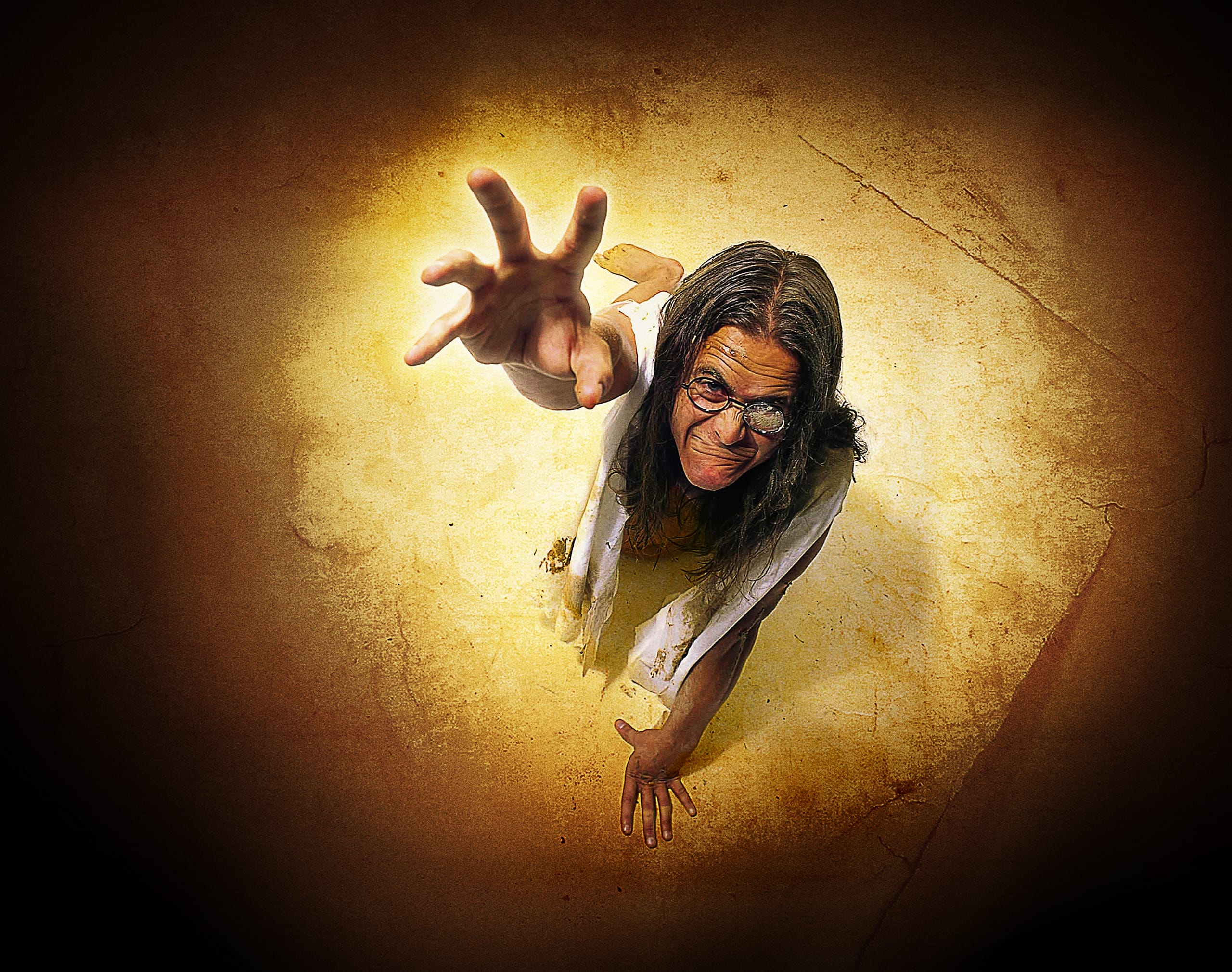 A parched person with long hair and glasses, wearing a tattered white shirt, is dramatically reaching out with one hand towards the camera, surrounded by a golden glow with a cracked, textured background, suggesting they are emerging from the ground. The expression is intense, filled with urgency.