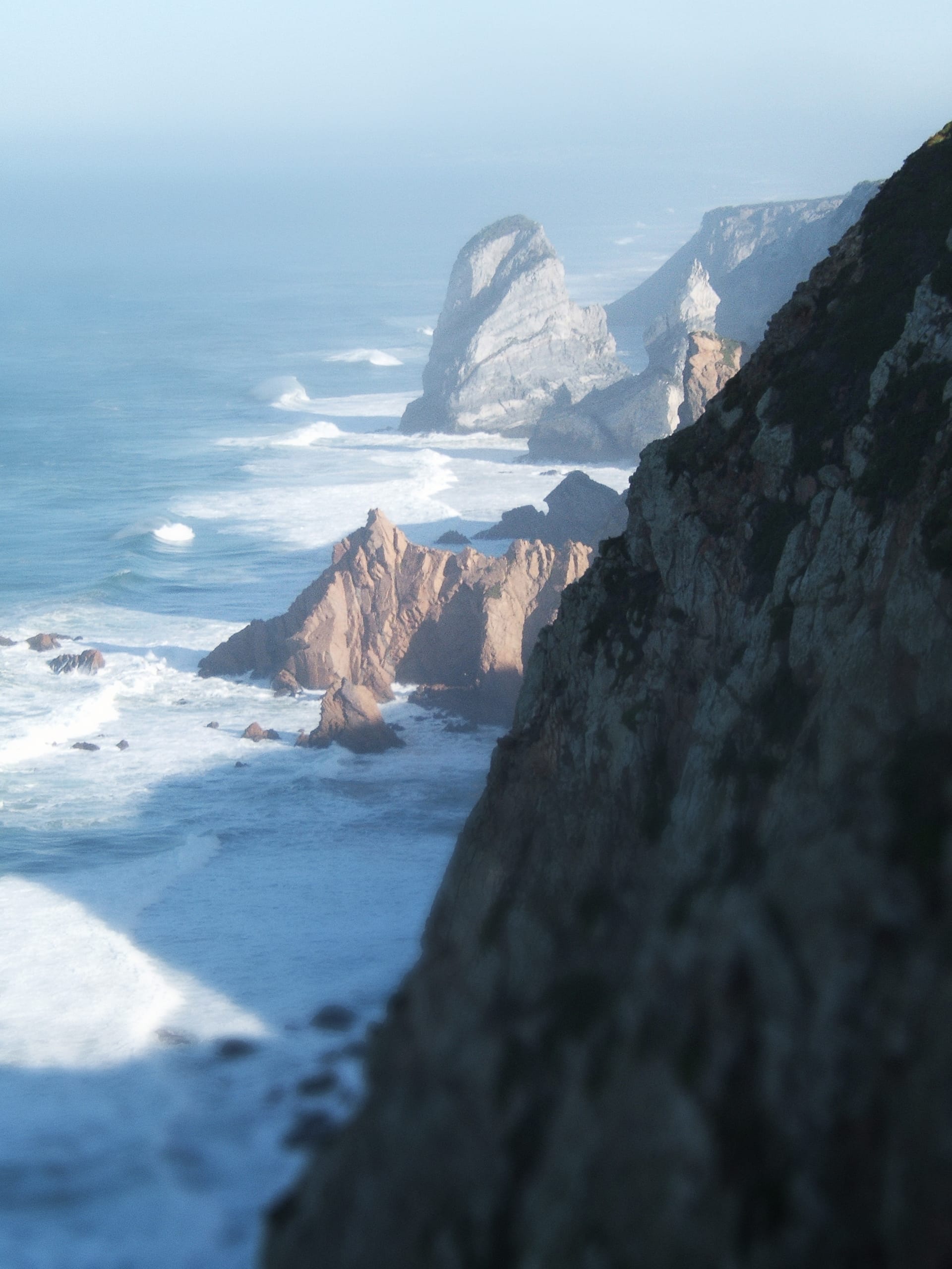 Steep, rugged cliffs with jagged rock formations extend into the ocean, where blue waves crash against the shore. The misty horizon blends sea and sky along the Portuguese Coastline, with layers of cliffs receding into the distance. The scene is bathed in a soft, diffused light.
