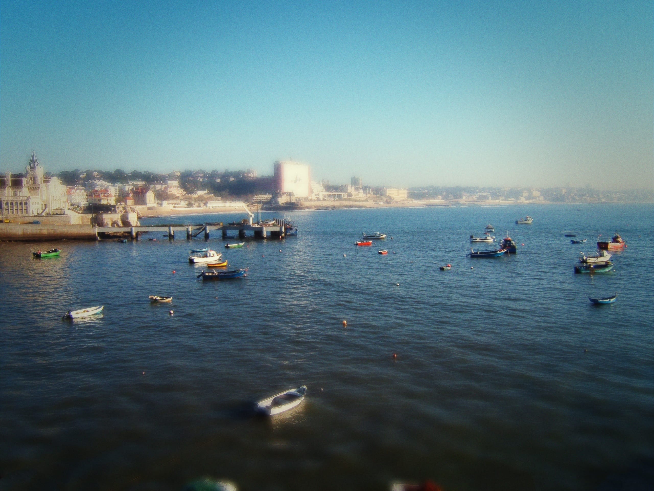 The image displays a serene Portuguese coastal scene with numerous small boats floated on calm waters. A pier extends into the harbor on the left side, leading to a town with various buildings. In the background, a slight haze covers the horizon, blending the sky and distant landscape.