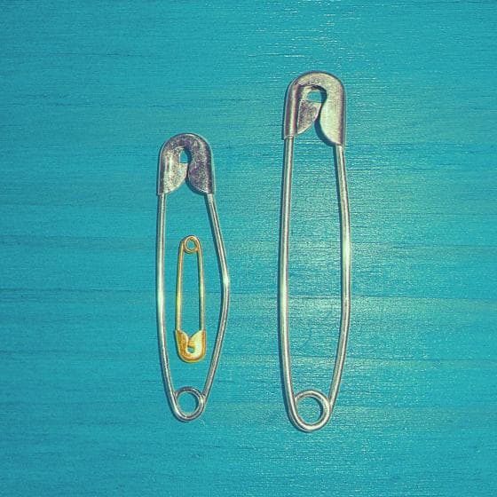 Two silver safety pins, one large and one small, lie parallel on a turquoise wooden surface. Inside the small silver pin, a tiny gold safety pin is nested. The vibrant blue background contrasts with the metallic sheen of the safety pins, creating a charming and subtle pregnancy announcement.