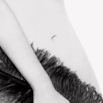 A black and white close-up image of a person's midsection, showing the left side of their torso and part of their lower abs. The texture of a large feather appears to overlap the lower right area of the image, blending seamlessly with the skin. This striking piece is part of the Presence Series. The background is white.