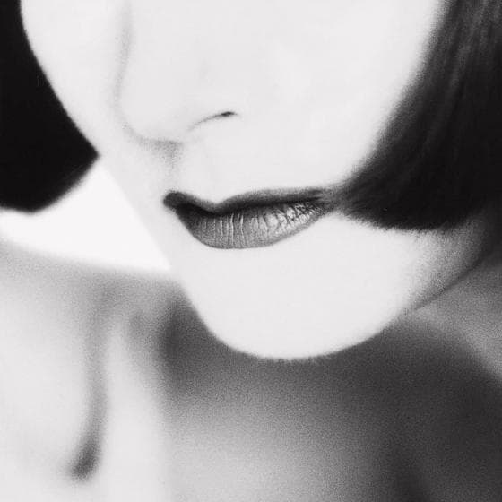 A high-contrast black-and-white photograph from the Presence Series focuses on the lower half of a person's face. The person has closely cropped dark hair and wears dark lipstick, providing a striking contrast against their pale skin. Only the lips, part of the nose, chin, and shoulder are visible, creating a dramatic and abstract appearance. 10/10 for visual impact!