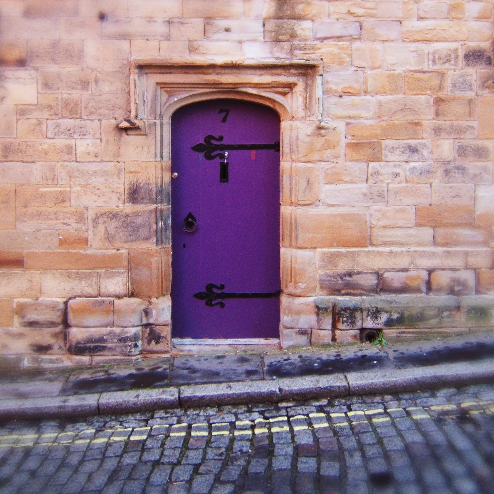 A vibrant purple door, labeled with the number 7, set in a rustic stone wall with a mix of tan and beige bricks, adds a unique touch to home decor. The door features large, ornate black hinges and a black knocker. A cobblestone street with yellow lines lies in front of the entrance, showing signs of age and weathering.