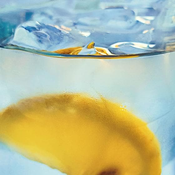Close-up image of a glass of iced water with a lemon slice. The top part of the image shows melting ice cubes, slightly distorted by the water's surface. The bottom part of the image clearly displays the lemon slice, with its bright yellow color contrasting against the clear water.