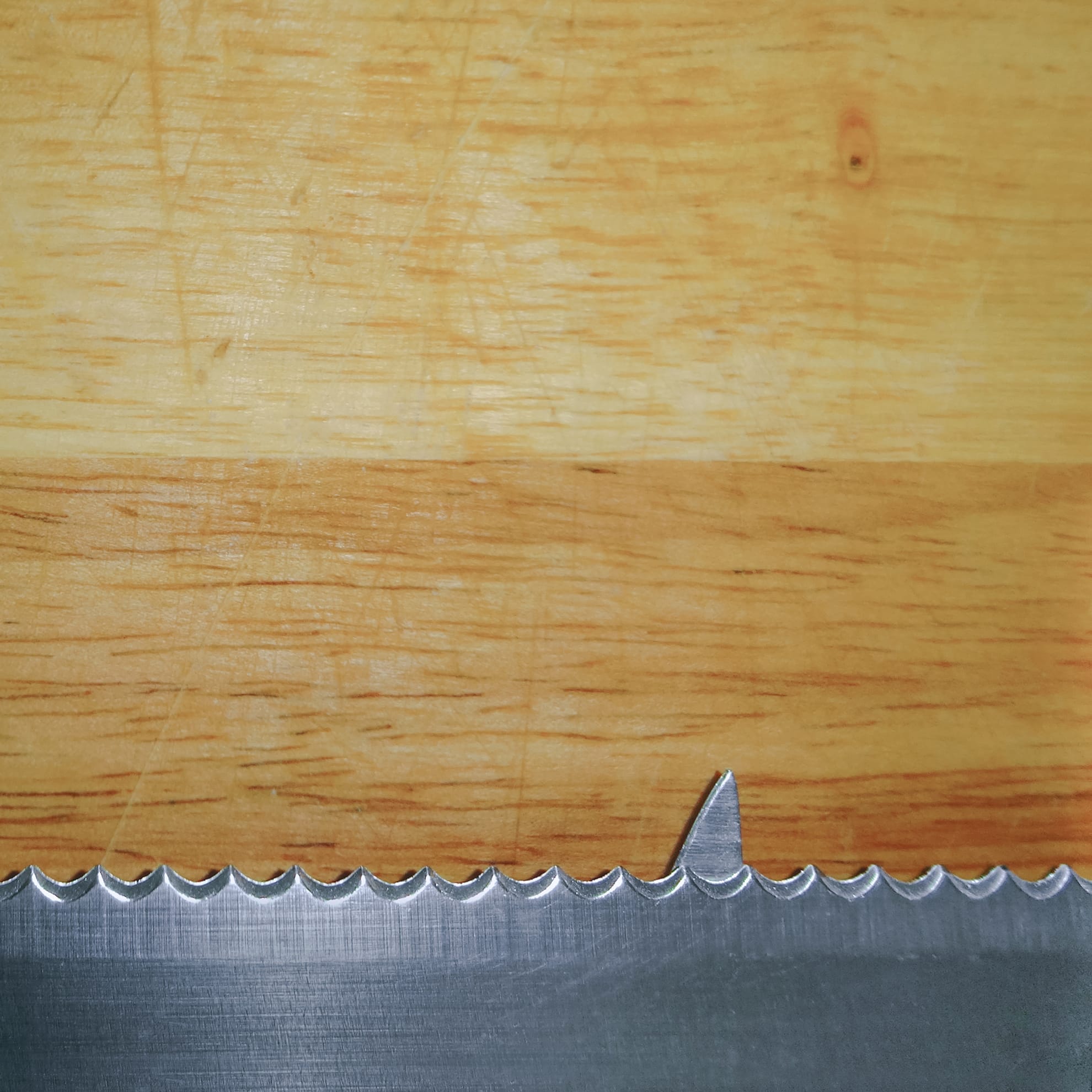 Close-up of a serrated knife blade resting on a wooden cutting board. The blade's sharp, jagged edge contrasts against the smooth, light-colored wood with visible grain patterns. The image focuses on the texture and detail of both the metal blade and the wooden surface.