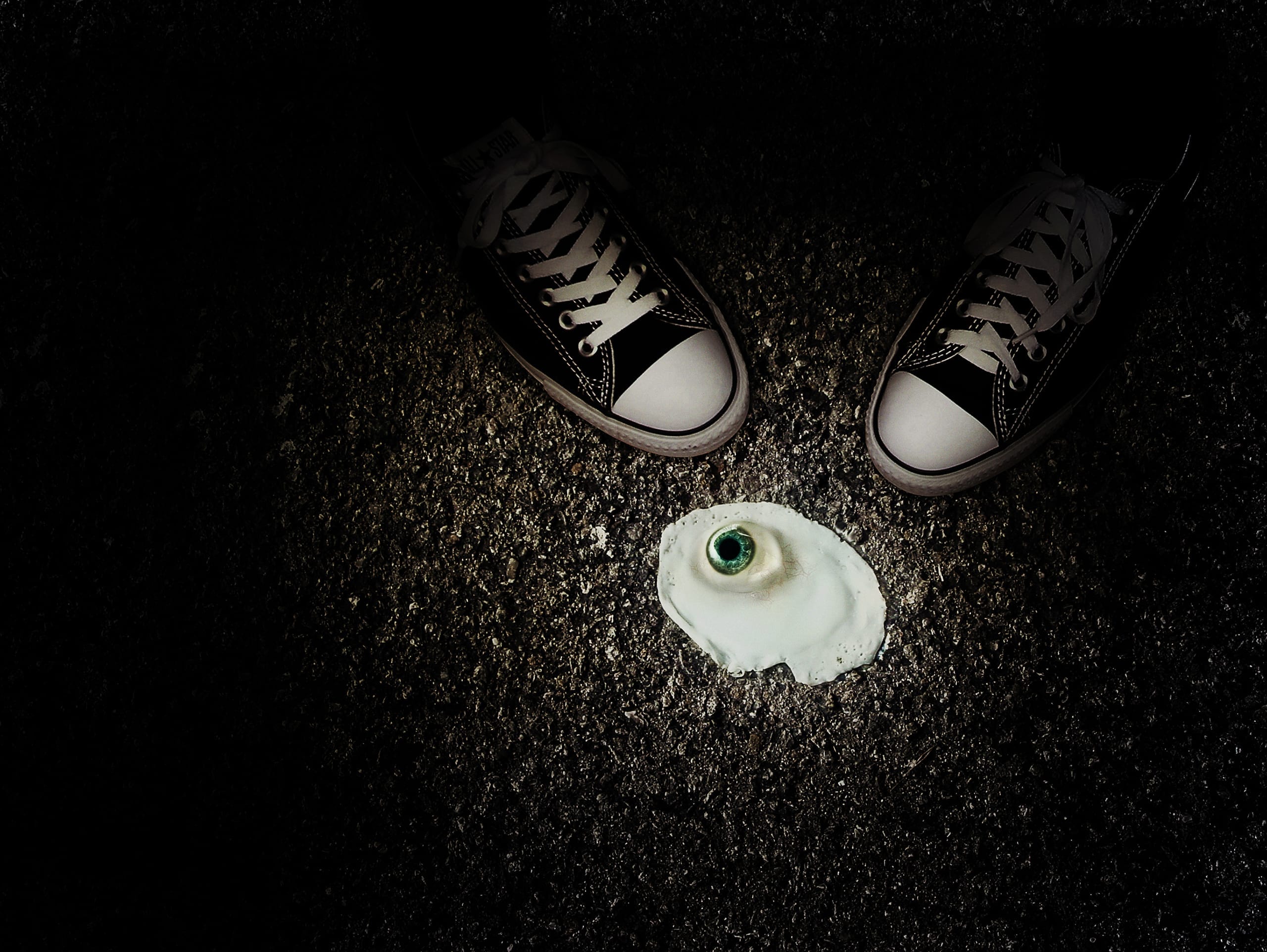 A pair of black and white sneakers stand on dark pavement, framing a fried egg with a surreal twist—a realistic green eye embedded in the yolk, gazing upwards. The surrounding area is dimly lit, emphasizing the eerie and unusual subject, as if hinting at some mysterious eye cooking.