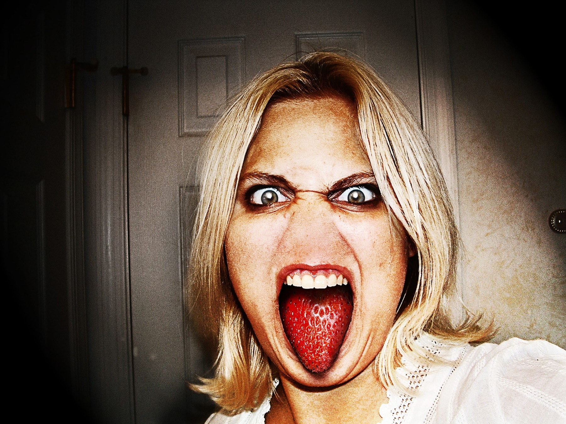 A person with light blonde hair is making a humorous face, showing wide eyes and sticking out their tongue, which is digitally manipulated to resemble a large strawberry. They are indoors, with a closed door and part of a wall visible in the background. The image lighting is slightly dark, as if taken during Strawberry Rehab.