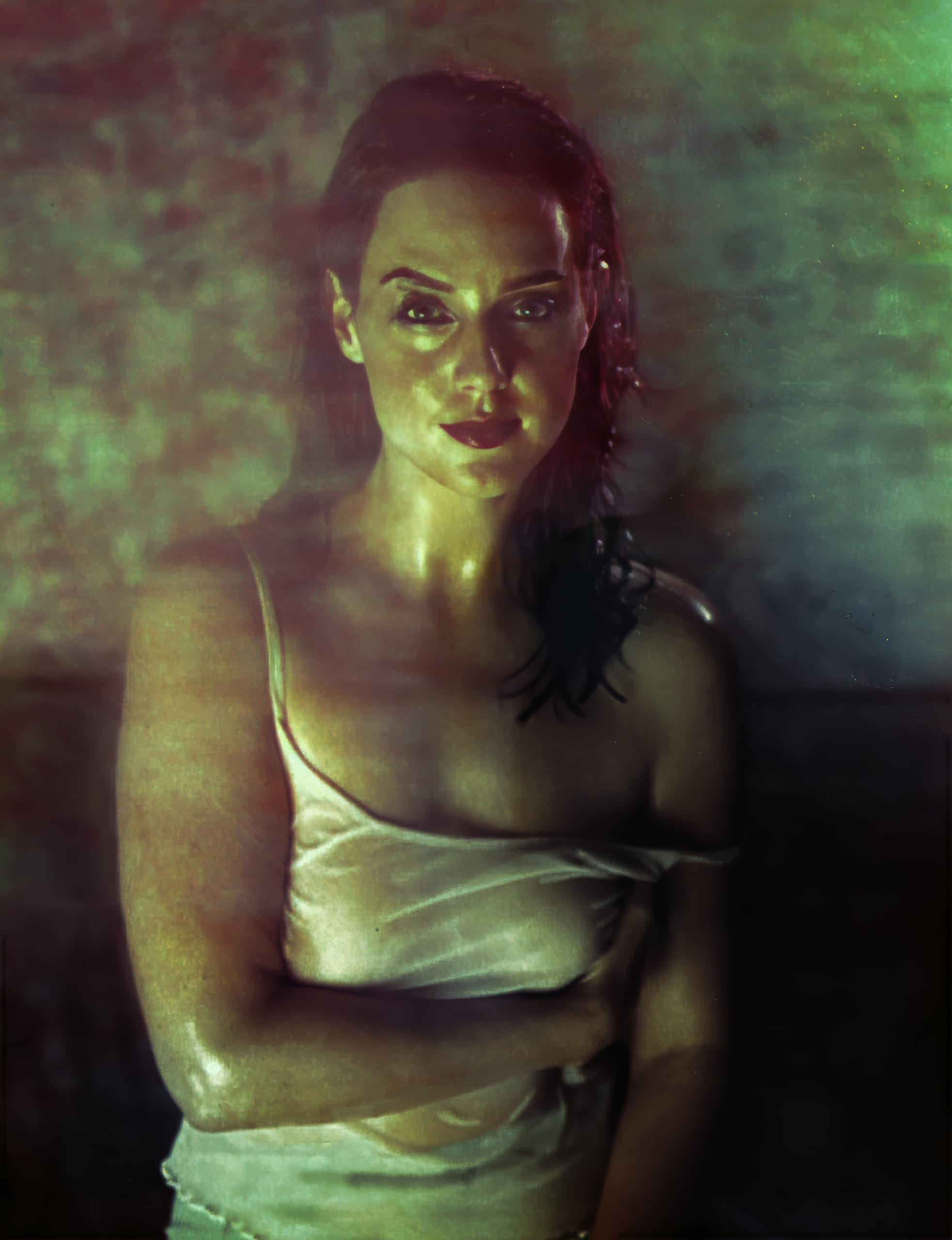 A woman with wet hair and dewy skin stands in dim, greenish lighting. She wears a simple white camisole, with one strap slipping off her shoulder. Her expression is serious, and the image has a moody, atmospheric feel with soft focus around the edges.