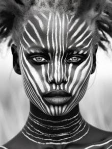 A person with intricate white line patterns painted on their dark skin. The lines radiate symmetrically from the nose across the face and continue down the neck. Their hair is styled in textured sections. The background is softly blurred, drawing focus to the detailed face art.