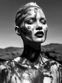 A black and white photo of a person with short hair, eyes closed, and head slightly tilted back. Their face and upper body are artistically covered in dark, paint-like streaks. The background features blurred, out-of-focus hills. The lighting creates strong contrasts and shadows.