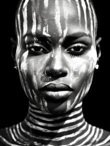 A grayscale portrait of a person with white tribal paint adorning their face and neck. The paint consists of vertical and horizontal stripes, creating a striking contrast against their dark skin. The expression is calm and the background is solid black, highlighting their features.