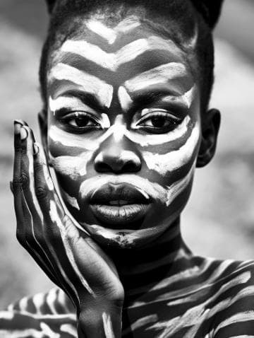 A black and white photograph of a person with intricate face and body paint. White patterns are painted on their face and hand, resembling abstract or animal-inspired designs. The person's expression is calm, with their hand gently touching their cheek. The background is blurred.