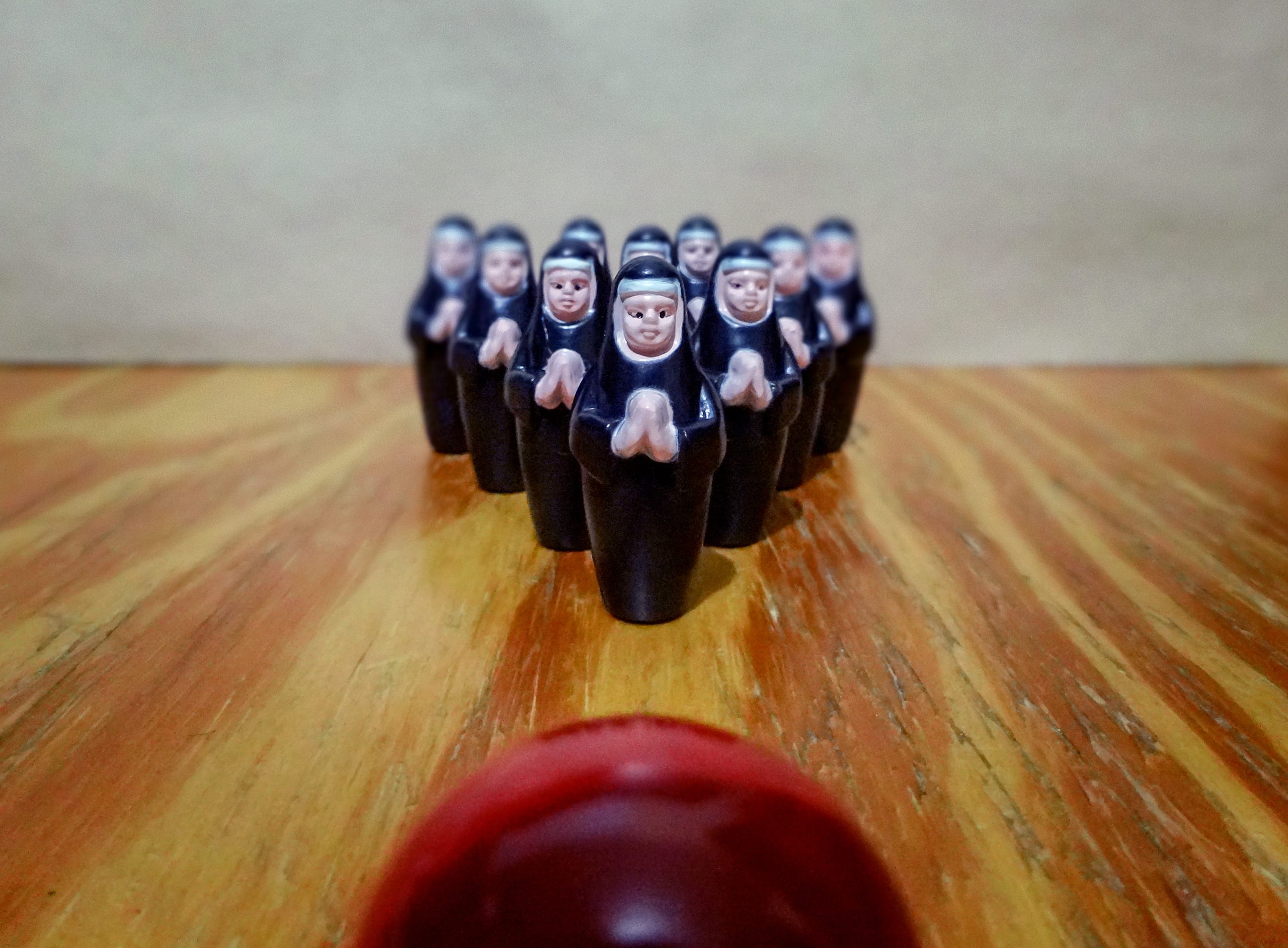 A row of miniature nun figurines are positioned like bowling pins on a wooden surface. They are dressed in black and white habits, with hands together in prayer. A red bowling ball is in the foreground, slightly blurred, as if rolling towards the nuns.