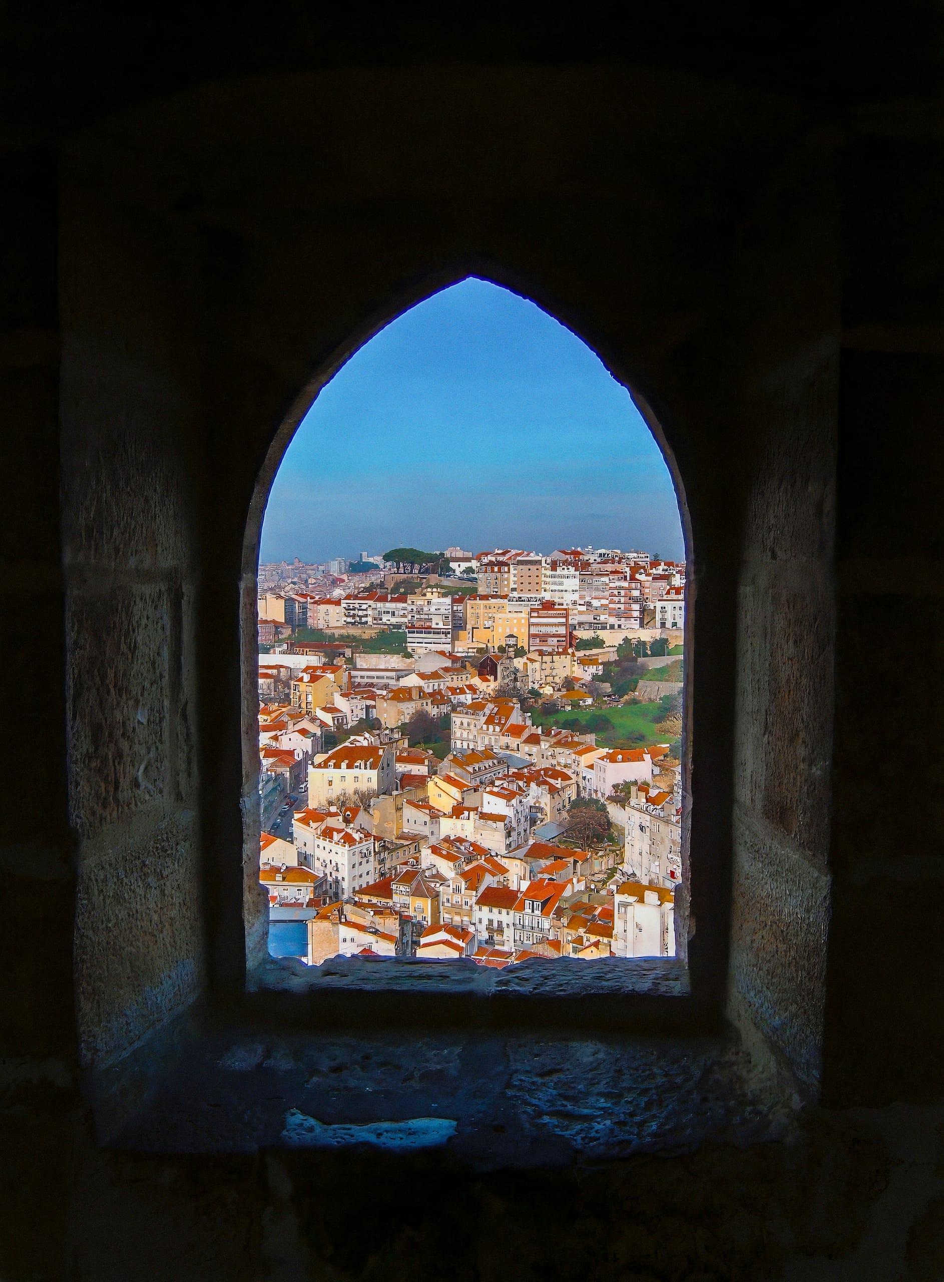 View of a Lisbon cityscape framed by a stone arched window, reminiscent of Belem Tower. The scene features red-tiled rooftops and varied buildings set against greenery and a clear blue sky. The window crafts a vignette effect, drawing focus to the picturesque urban landscape outside.