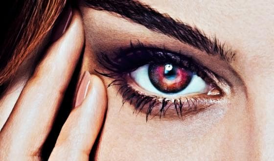 Close-up image of a person's eye. The iris is a vivid, striking red with colorful reflections, and the eyelashes are long and dark. The eyebrow is well-defined, and the person's hand gently touches the side of their face near the eye, showcasing neatly manicured nails—a mesmerizing eye study of unique visions.