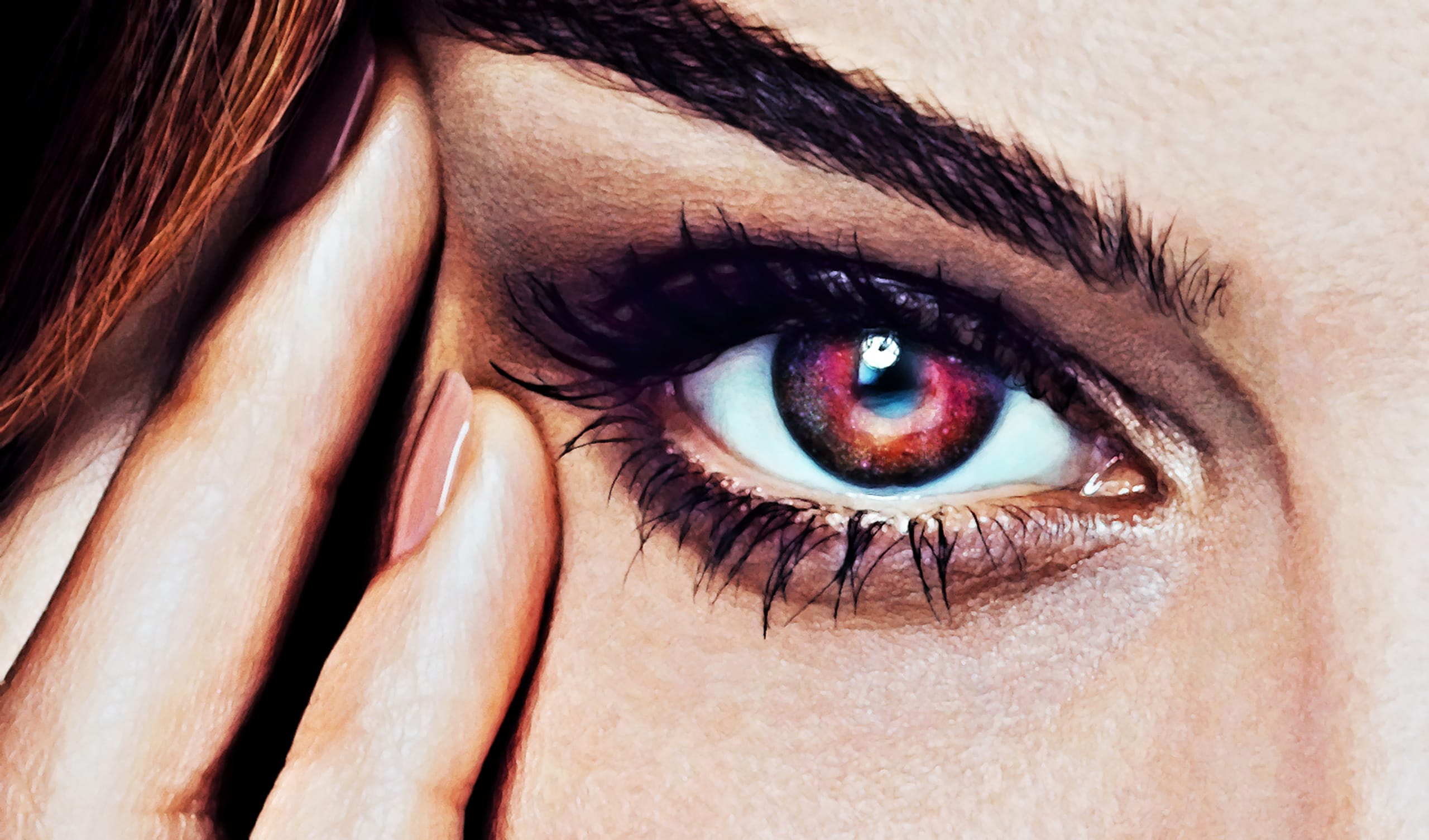 Close-up image of a person's eye. The iris is a vivid, striking red with colorful reflections, and the eyelashes are long and dark. The eyebrow is well-defined, and the person's hand gently touches the side of their face near the eye, showcasing neatly manicured nails—a mesmerizing eye study of unique visions.