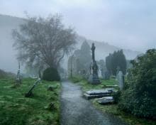 A misty cemetery with a winding gravel path leading through. Weathered gravestones and a tall monument stand amidst lush green grass and leafless trees. Fog envelops the scene, partially obscuring distant hills and creating an eerie yet serene atmosphere.