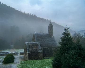A small, ancient stone church with a tall spire sits surrounded by misty, forested hills. The ground is lightly dusted with frost or snow, and a path leads to the church. Dense evergreen trees and fog add to the serene and mysterious atmosphere of the scene.