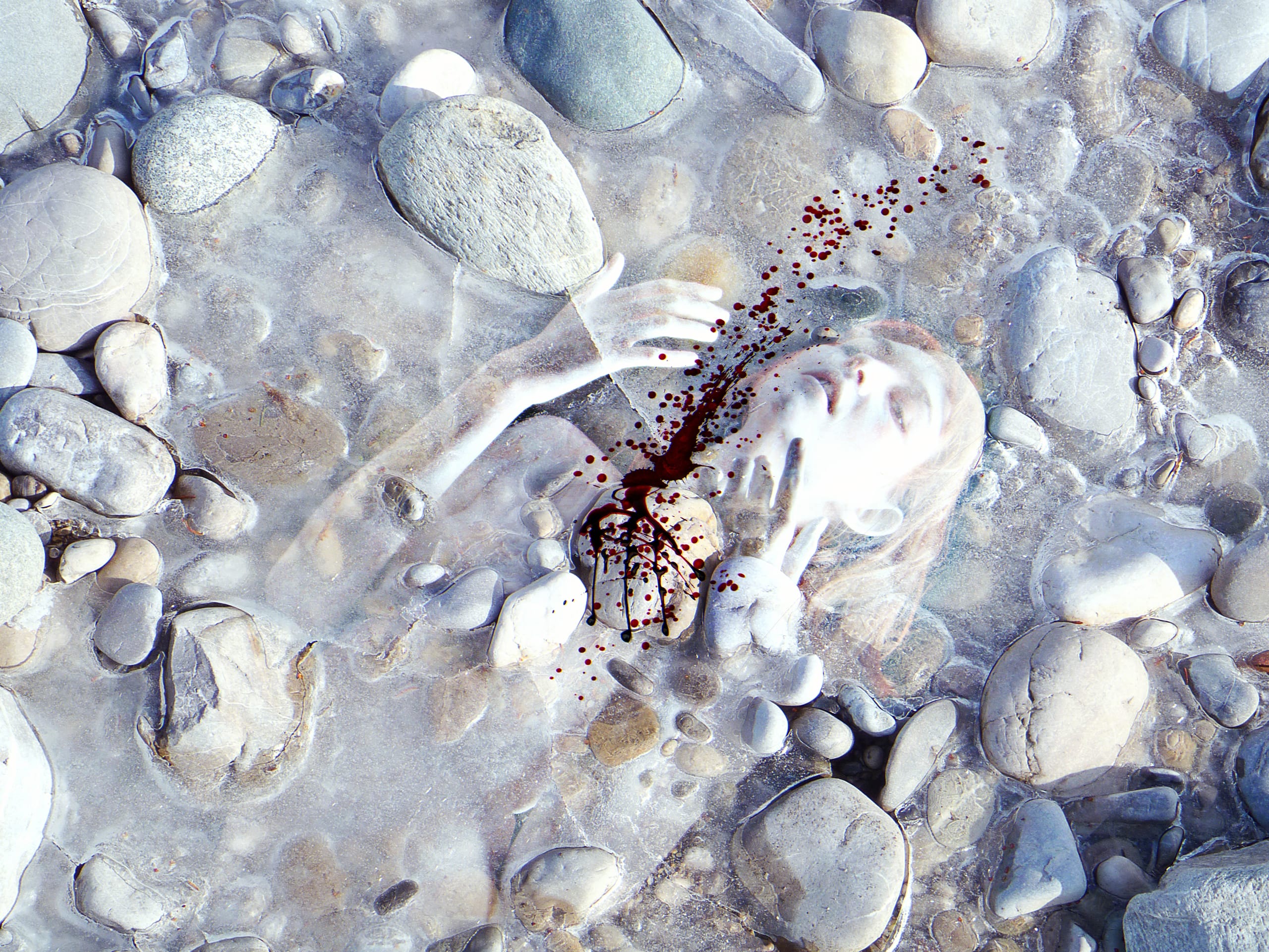 A surreal image of a person seemingly submerged beneath a rocky surface. Their face and hands are visible through the stones, with some rocks partially obscuring them. Blood splatter is present near their left hand and face, creating a dramatic and eerie atmosphere.