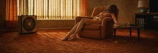 Woman in an orange chair reaches for snacks, with sunlight and nature reflected through blinds onto the carpet and tabletop.